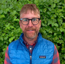 Matt Madsen Man with glasses and a blue vest smiling in front of greenery