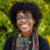 Woman with glasses standing in front of bushes smiling 